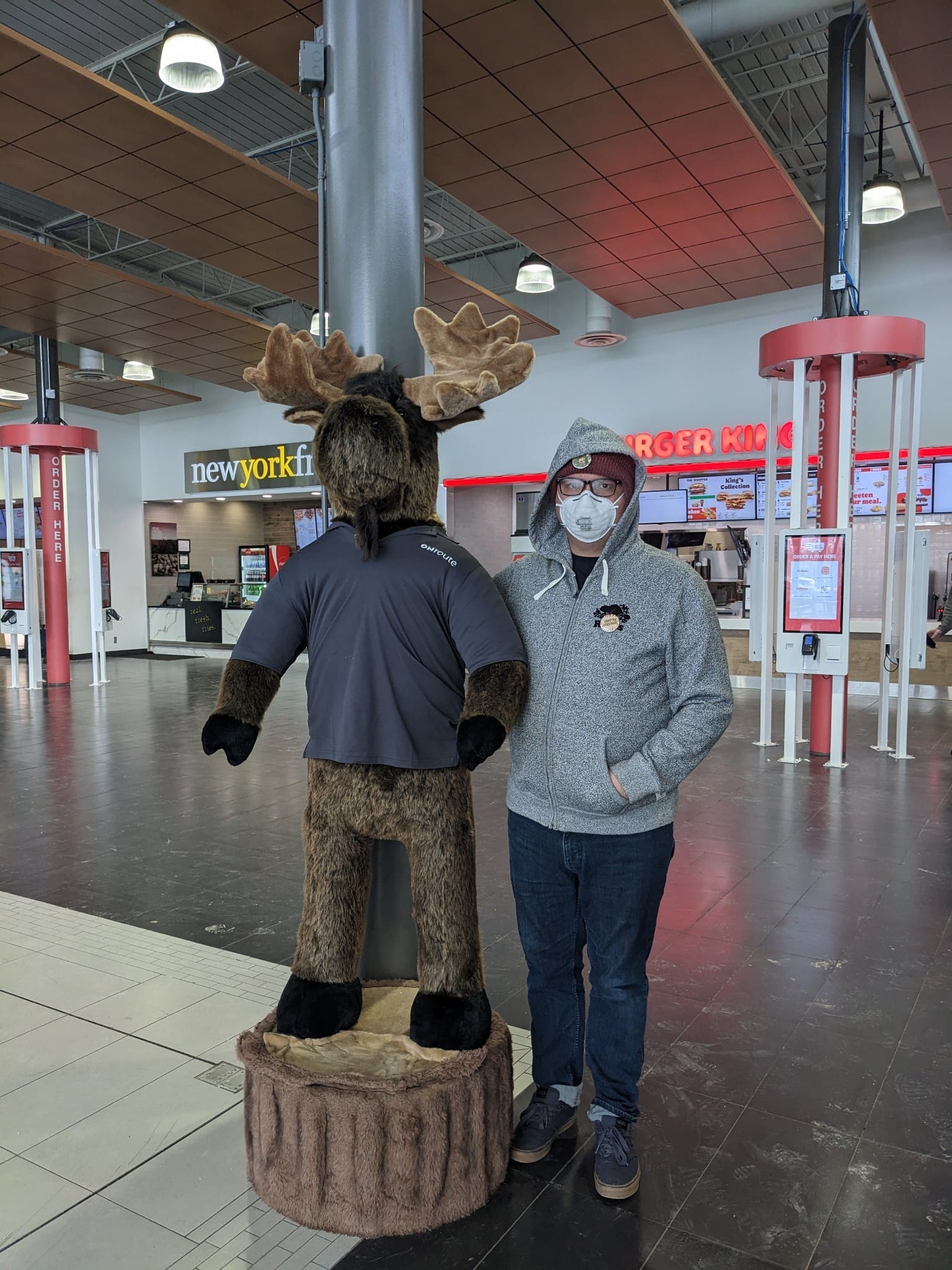 A man in a toque, N95 mask and grey sweater next to a plush moose his height in a food court